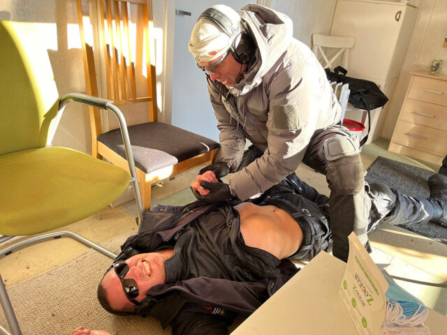 En mann ligger på gulvet under øvelse. En annen mann forsøker hjelpe han.