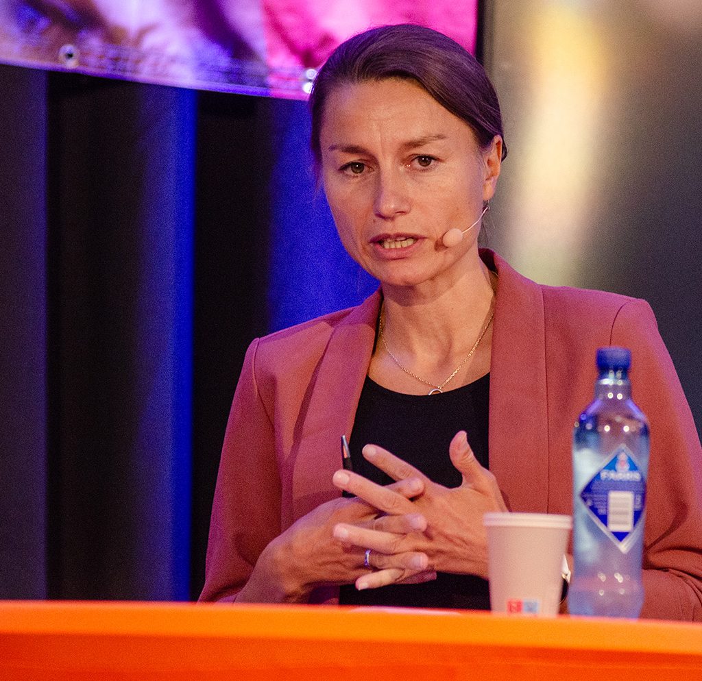 Foto av kvinne i rød blaser. Hun står på en scene.