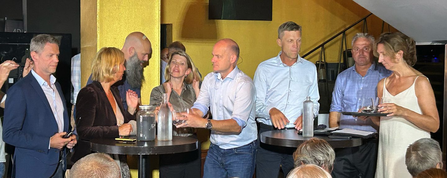 En gruppe ansatte fra PST står samlet rundt et par bord på en scene.