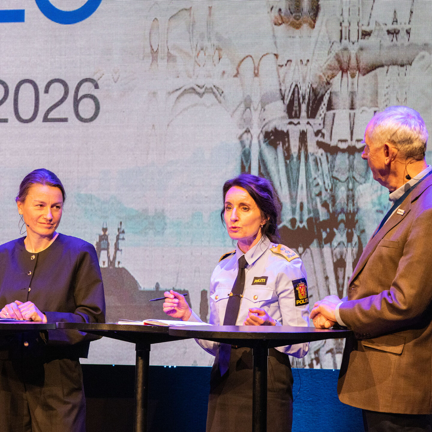 Tre personer på en scene. De snakker med hverandre. Foto.