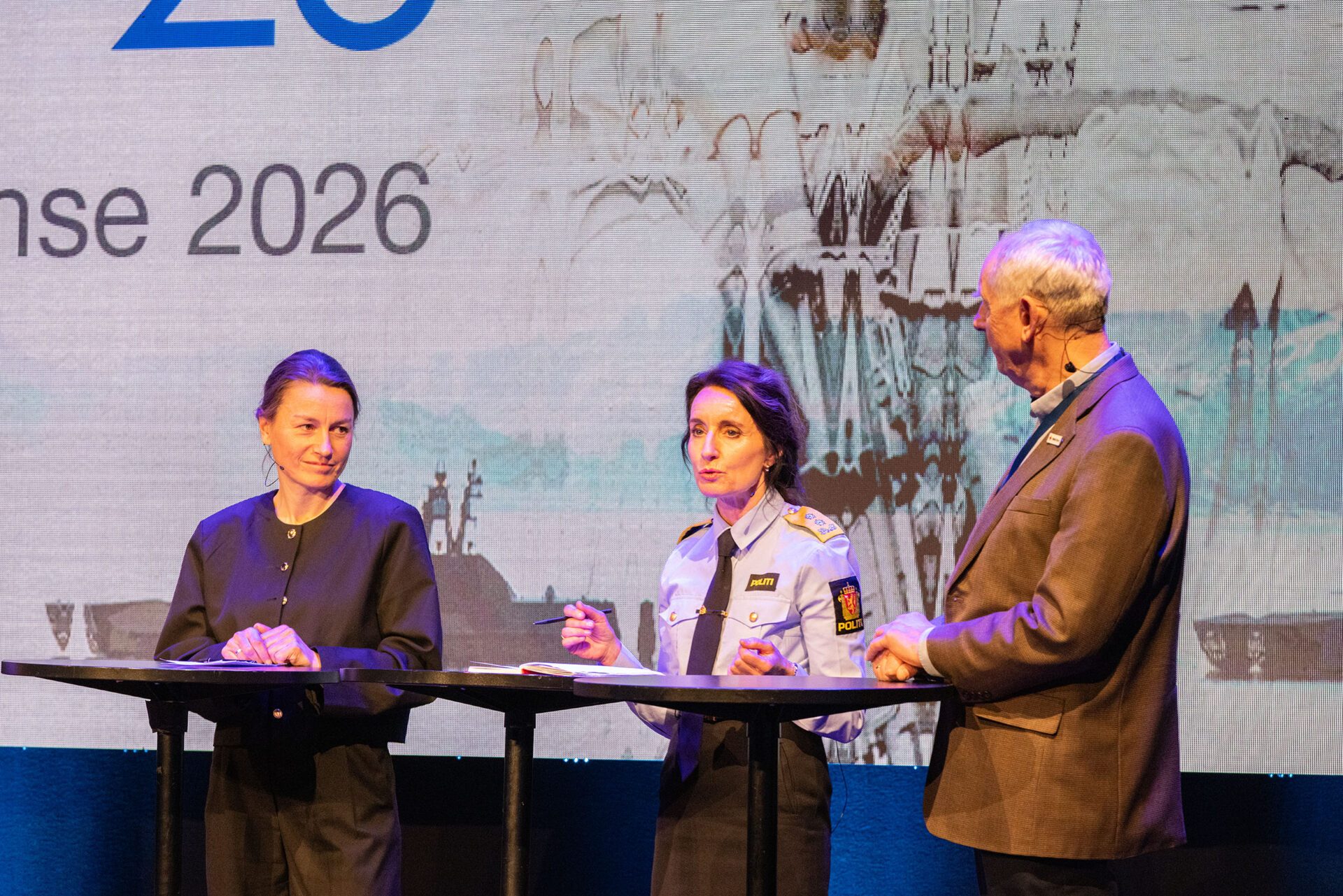 Tre personer på en scene. De snakker med hverandre. Foto.