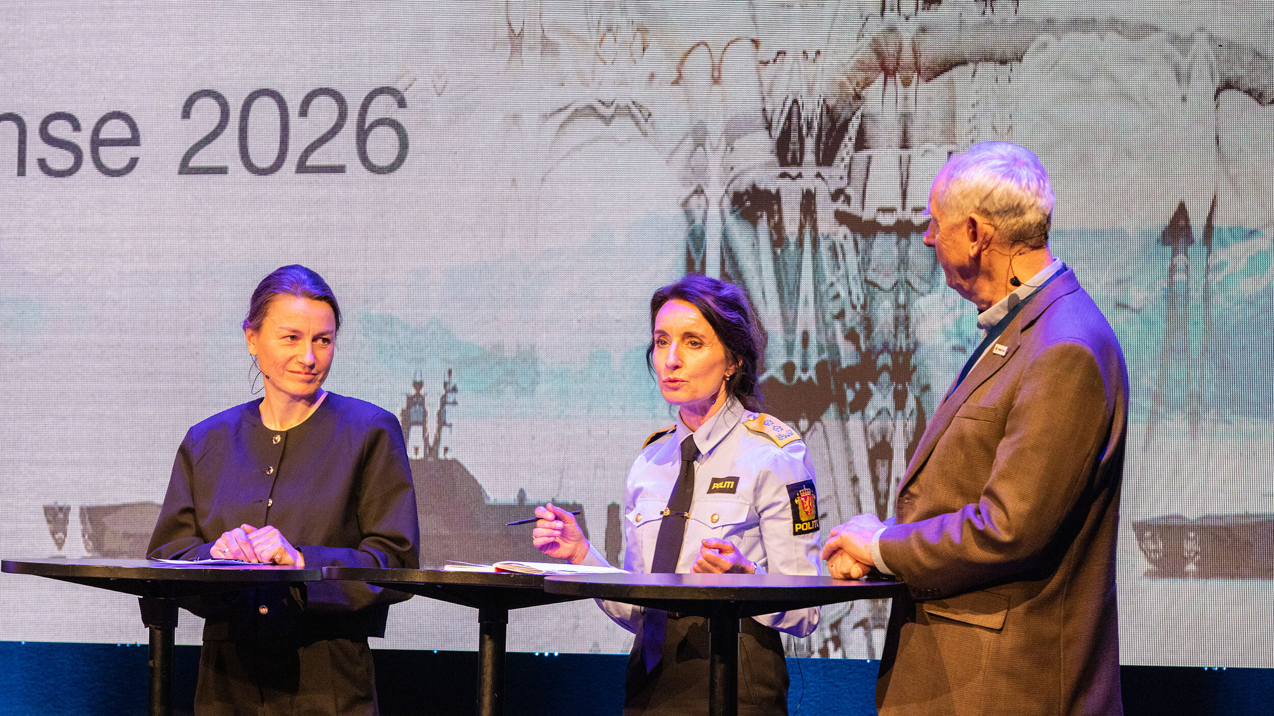 Tre personer på en scene. De snakker med hverandre. Foto.