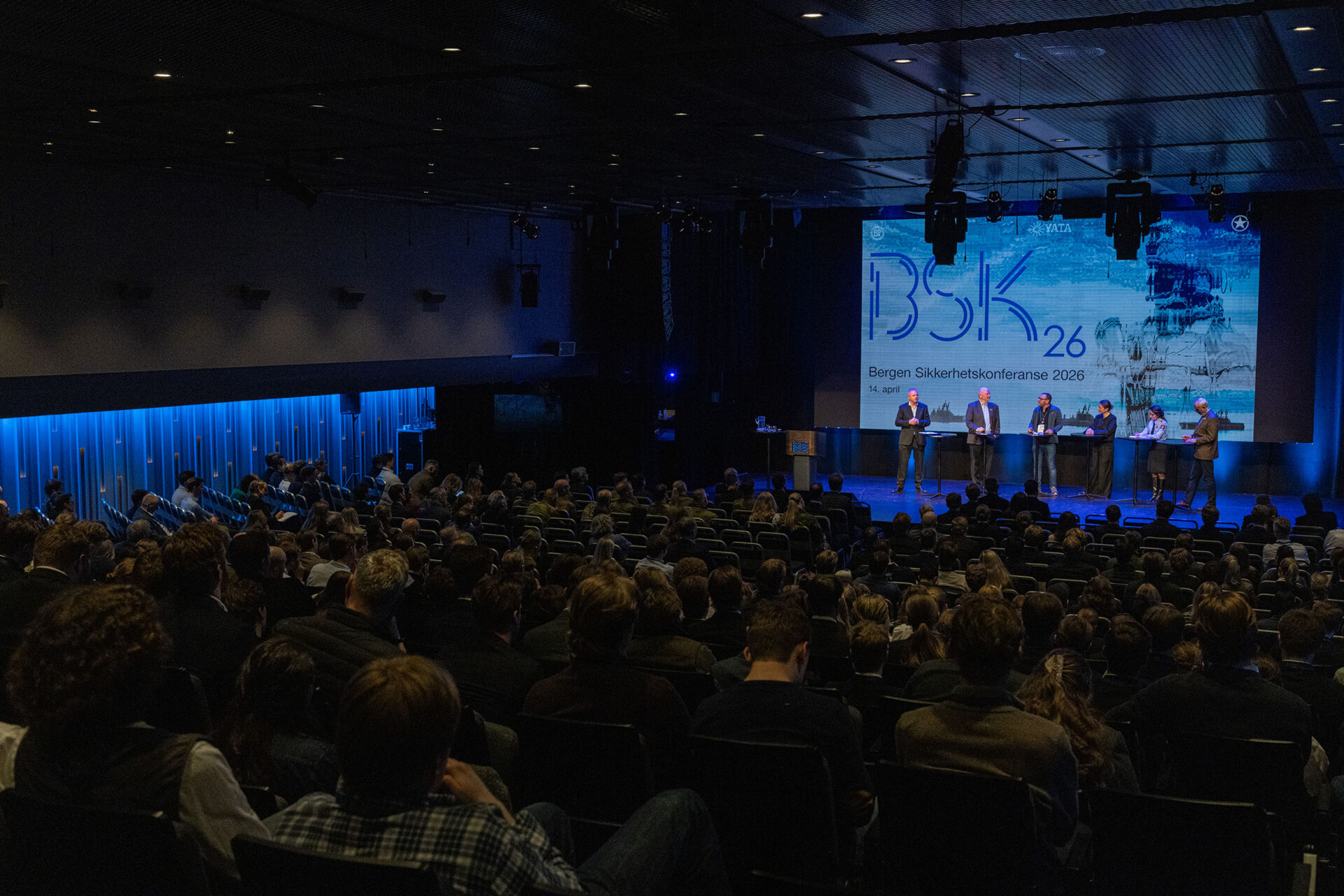 Mørklagt konferansesal med mange tilskuere som ser på et panel som står foran en stor skjerm. Foto.