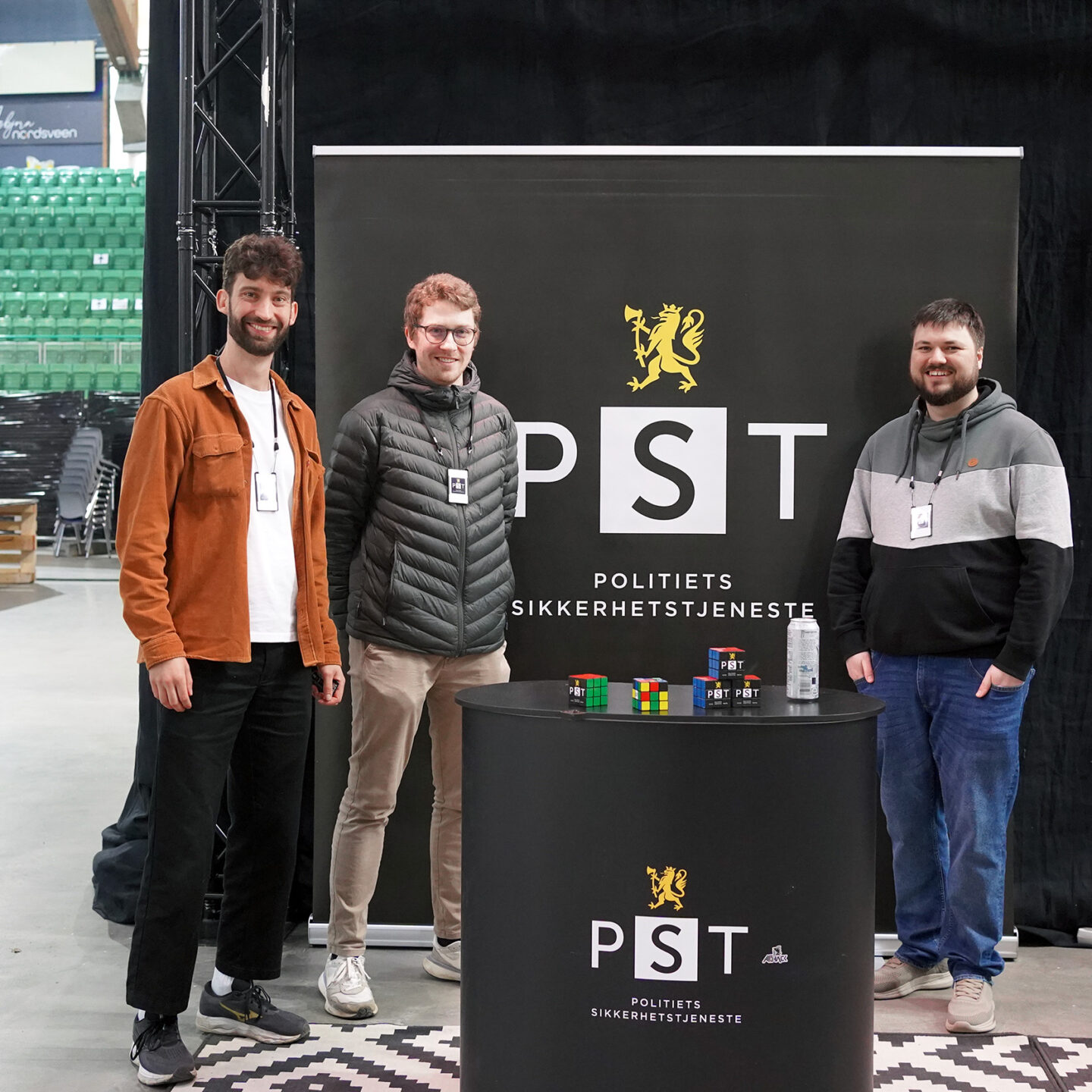 Tre teknologer i PST står foran en mørk bakvegg med PST-logo. Foto.