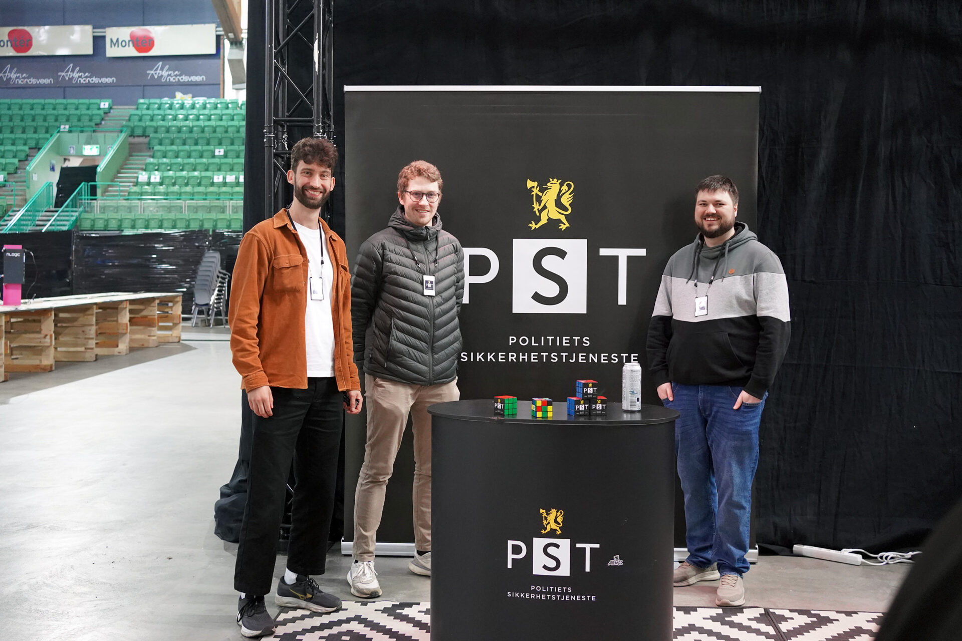 Tre teknologer i PST står foran en mørk bakvegg med PST-logo. Foto.