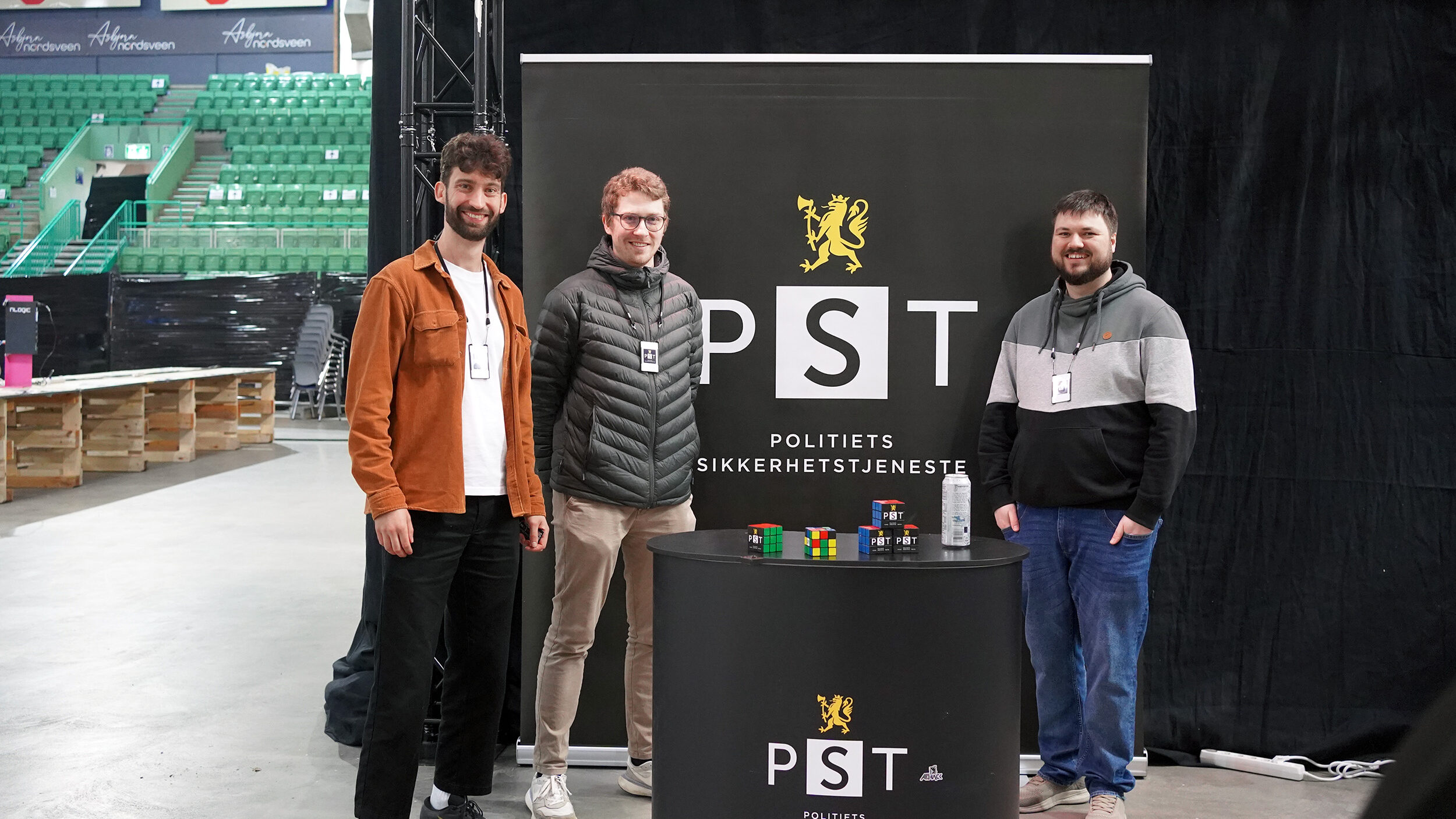 Tre teknologer i PST står foran en mørk bakvegg med PST-logo. Foto.