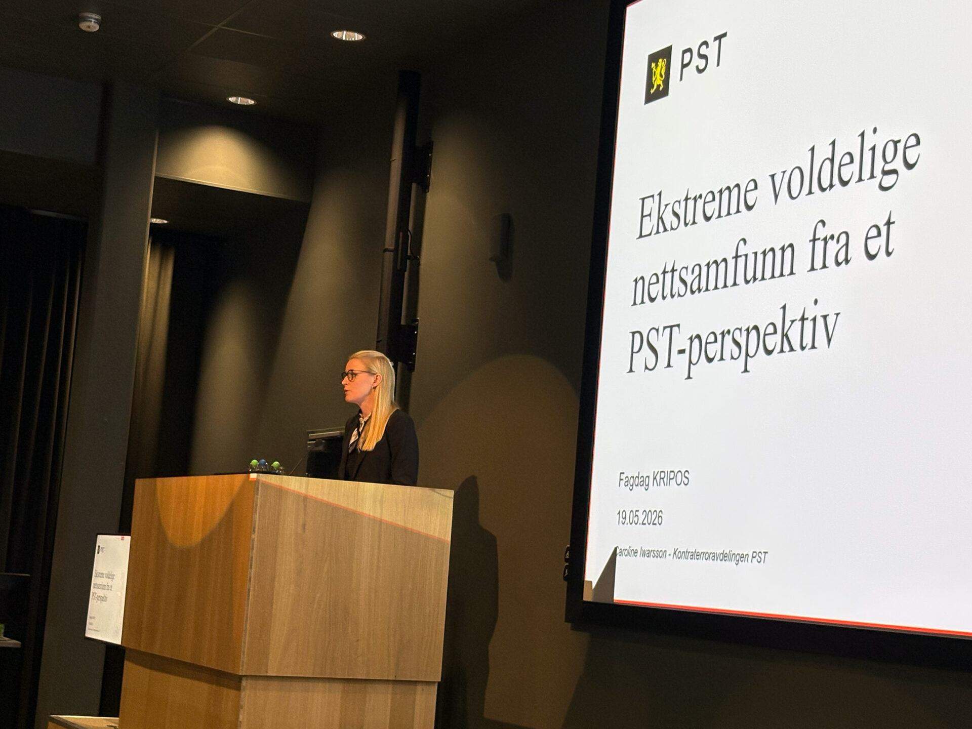 PST-ansatt står ved en talerstol. Snakker til befolkingen. Bak henne er en skjerm som viser en presentasjon. Foto